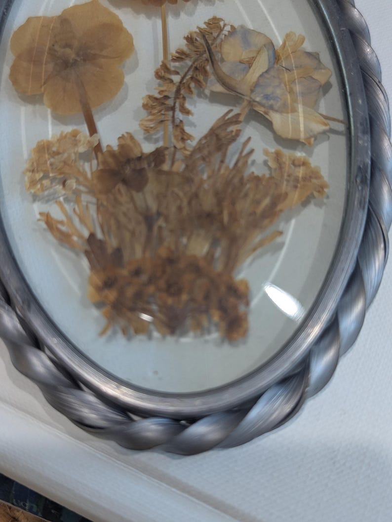 May include: An oval-shaped framed pressed flower arrangement. The frame is silver with a twisted rope design. The dried flowers are various shades of brown and tan, with some hints of blue. The arrangement is encased in glass.