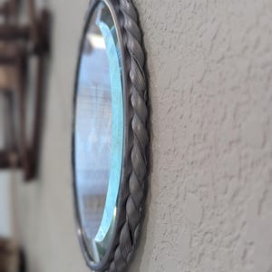 May include: Oval mirror with a gray braided frame hanging on a textured wall. The mirror's frame has a woven rope design. The glass reflects the surrounding environment. The mirror is attached to the wall with a metal hook.