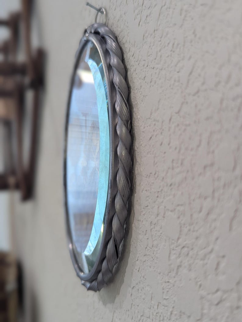 May include: Oval mirror with a gray braided frame hanging on a textured wall. The mirror's frame has a woven rope design. The glass reflects the surrounding environment. The mirror is attached to the wall with a metal hook.