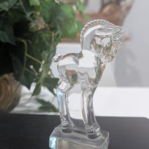 May include: A clear glass figurine of a horse standing on a rectangular base. The horse has a detailed mane and tail, and its body reflects light, creating a shimmering effect. The figurine is a decorative item.