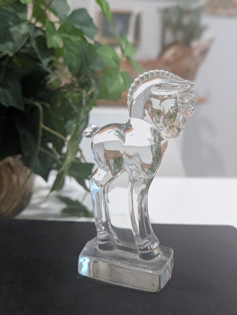 May include: A clear glass figurine of a horse standing on a rectangular base. The horse has a detailed mane and tail, and its body reflects light, creating a shimmering effect. The figurine is a decorative item.