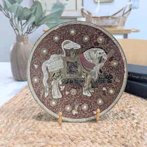 May include: A decorative, round, brass plate with a maroon background and an embossed design. The plate features a camel and horse with intricate detailing. The plate is displayed on a gold stand, showcasing its ornate craftsmanship.