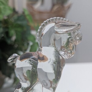 May include: A clear glass figurine of a donkey. The donkey is standing with its head turned to the side. The figurine has detailed features, including a mane and tail. The glass is transparent, allowing light to pass through.