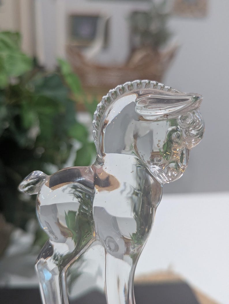 May include: A clear glass figurine of a donkey. The donkey is standing with its head turned to the side. The figurine has detailed features, including a mane and tail. The glass is transparent, allowing light to pass through.