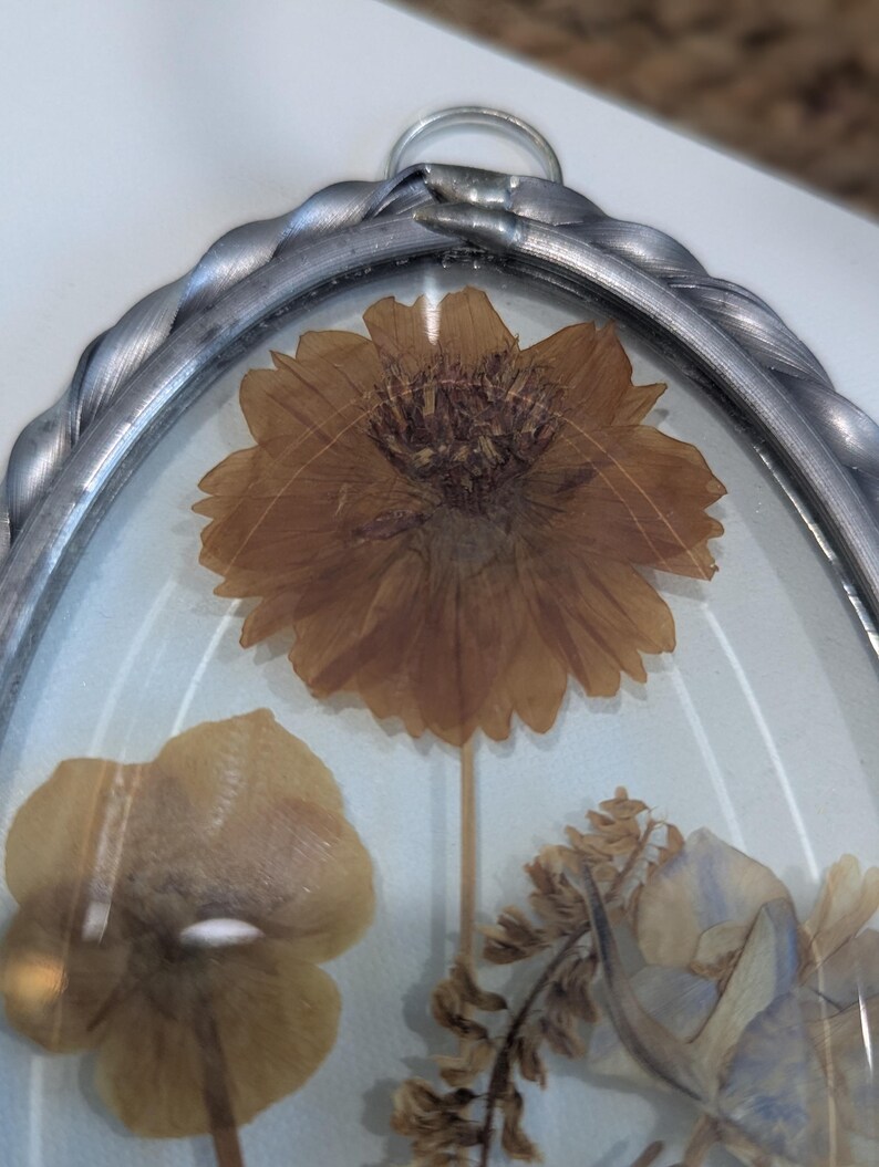 May include: An oval-shaped, pressed-flower art piece. The artwork features a large, dried, brown flower at the center, with other dried flowers in shades of yellow and brown. The flowers are encased in clear glass, framed by a silver-colored metal with a twisted rope design.