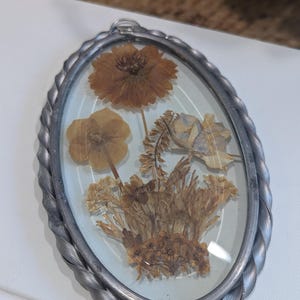 May include: Oval-shaped, pressed flower pendant with a silver-colored, braided metal frame. The pendant features dried flowers in shades of brown and tan, encased in clear glass. The frame has a small loop at the top for hanging.