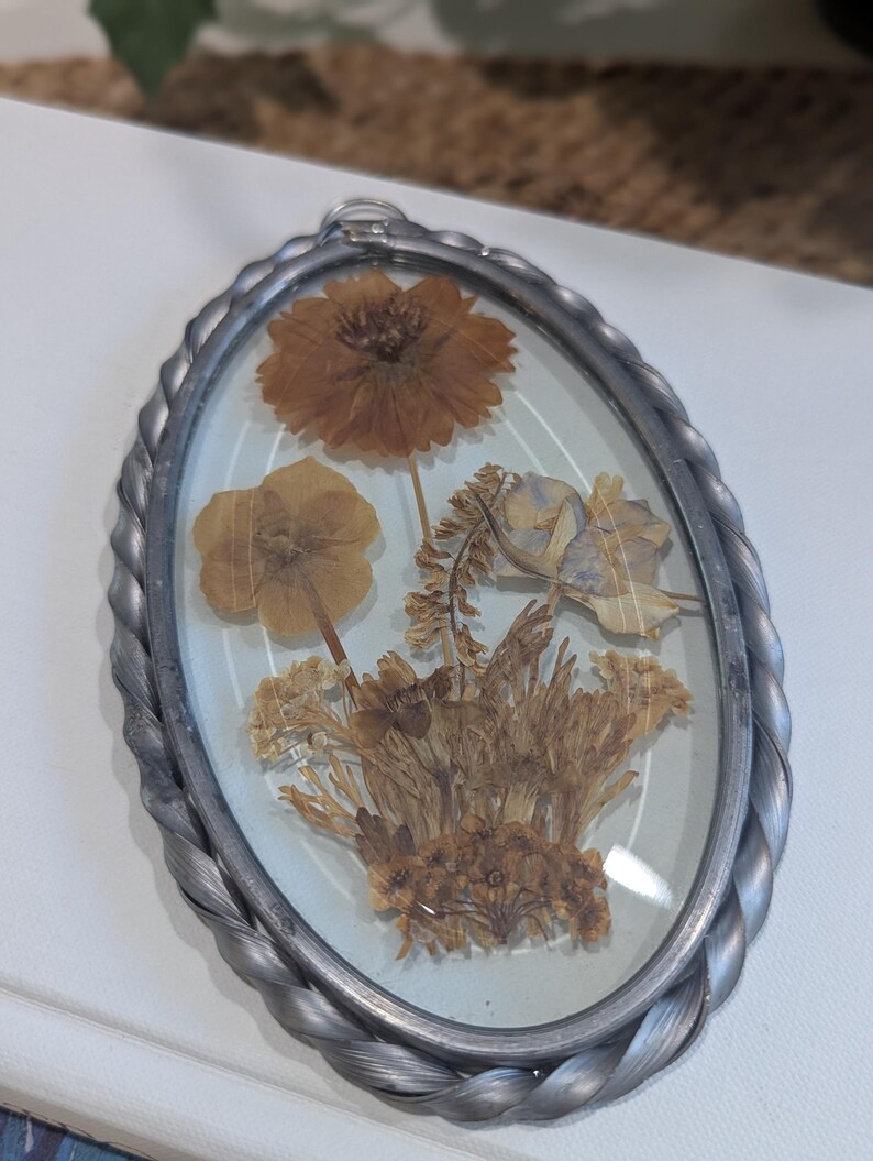 May include: Oval-shaped, pressed flower pendant with a silver-colored, braided metal frame. The pendant features dried flowers in shades of brown and tan, encased in clear glass. The frame has a small loop at the top for hanging.