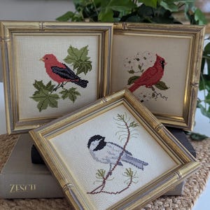 May include: Three square cross-stitch embroideries in gold bamboo-style frames. Each piece features a different bird: a red bird on green leaves, a red cardinal on white blossoms, and a black and white chickadee perched on a pine branch.