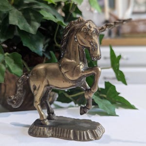 May include: A brass unicorn figurine with a detailed mane, horn, and raised front leg. The sculpture is mounted on a textured base and set against a backdrop of green foliage. The unicorn is a symbol of magic and fantasy.