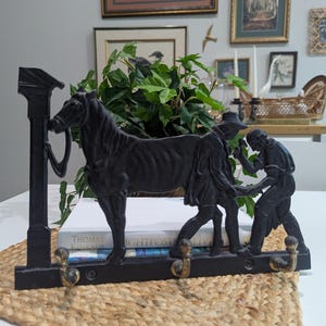 May include: Black cast iron wall hook featuring a horse, two figures, and three hooks. The horse is attached to a pillar. The figures appear to be working with the horse. The hooks have a decorative gold finish. The piece is on a woven mat.