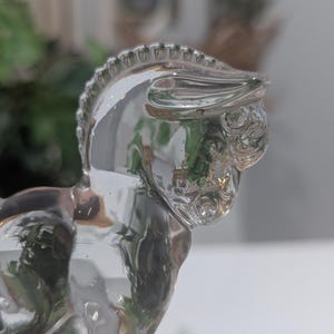 May include: A clear glass horse figurine. The horse is depicted in a side profile, with a detailed mane and facial features. The glass is transparent, reflecting the surrounding environment. The figurine is a decorative item.