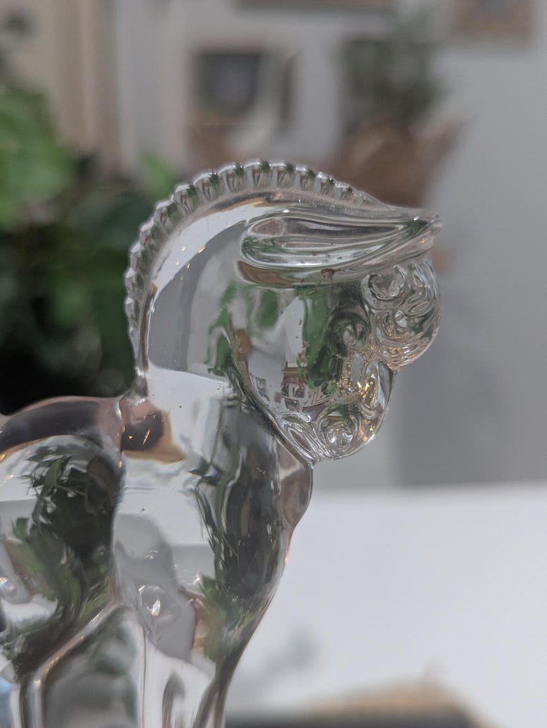 May include: A clear glass horse figurine. The horse is depicted in a side profile, with a detailed mane and facial features. The glass is transparent, reflecting the surrounding environment. The figurine is a decorative item.
