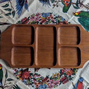 May include: A rectangular wooden serving tray with six compartments. The tray is made of a warm-toned wood and has a smooth finish. The tray is displayed on a floral patterned fabric with parrots.