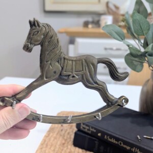 May include: A vintage-style brass rocking horse key holder. The horse is detailed with a saddle and mane, and the rocker base has hooks for keys. The piece has an aged, antique finish, and is a decorative home accent.