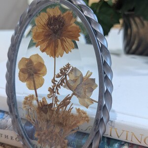 May include: Oval-shaped pressed flower art piece with a silver-colored braided metal frame. The glass displays dried flowers in shades of brown and tan. A small silver loop is at the top for hanging. The piece is resting on a stack of books.