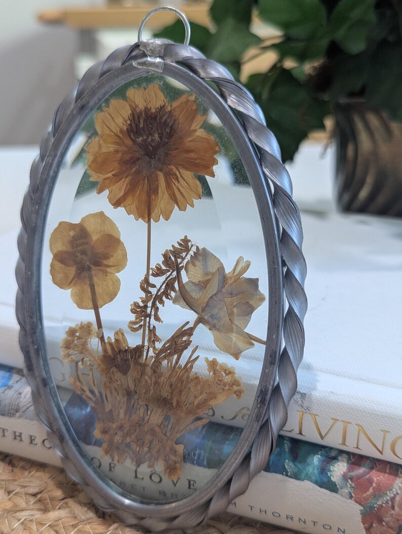 May include: Oval-shaped pressed flower art piece with a silver-colored braided metal frame. The glass displays dried flowers in shades of brown and tan. A small silver loop is at the top for hanging. The piece is resting on a stack of books.