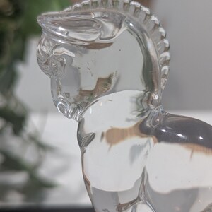 May include: A clear glass horse figurine. The horse is sculpted with detailed features, including a mane and defined musculature. The figurine is transparent, allowing light to pass through, and is set against a blurred background.