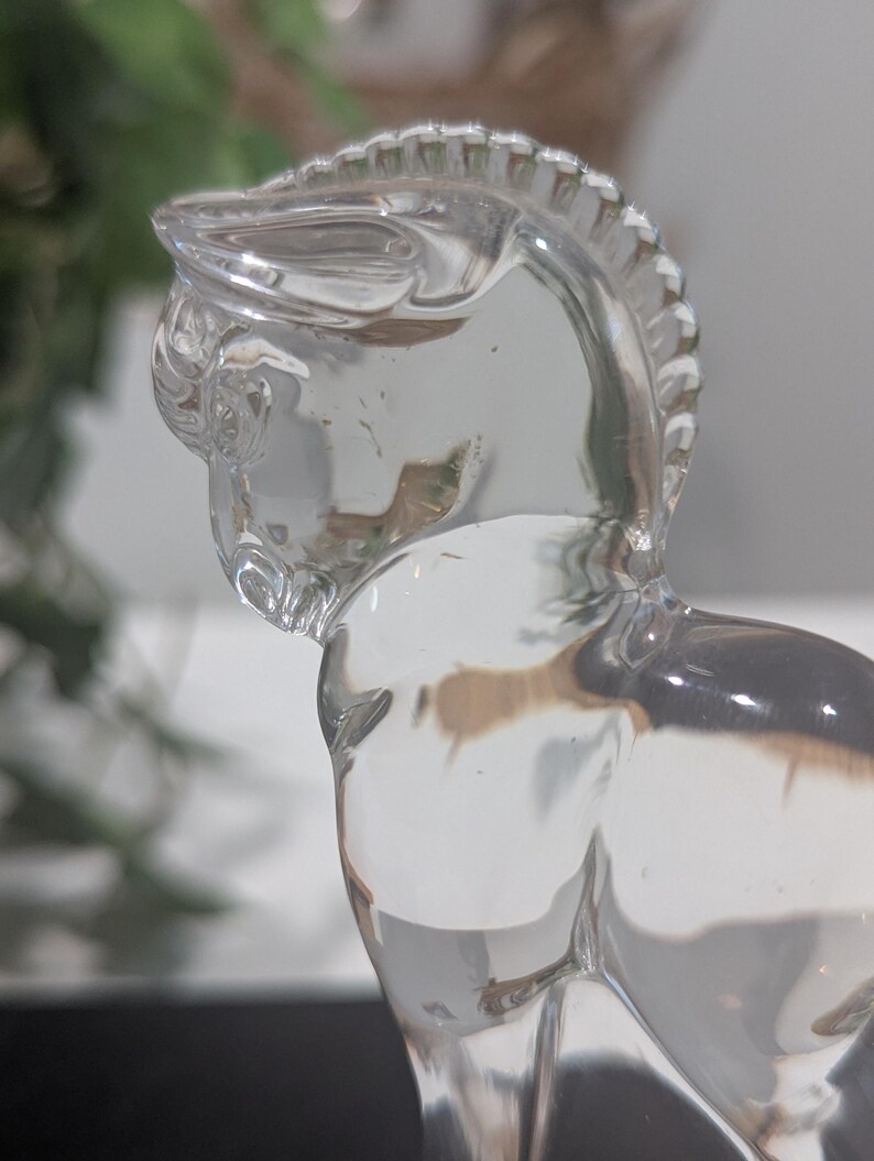 May include: A clear glass horse figurine. The horse is sculpted with detailed features, including a mane and defined musculature. The figurine is transparent, allowing light to pass through, and is set against a blurred background.
