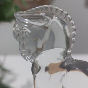 May include: A clear glass figurine of a horse, showcasing the head and upper body. The horse has a detailed mane and a smooth, reflective surface. The figurine is transparent, allowing light to pass through, and is set against a blurred background.