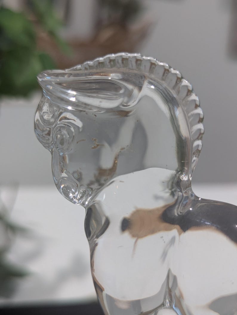 May include: A clear glass figurine of a horse, showcasing the head and upper body. The horse has a detailed mane and a smooth, reflective surface. The figurine is transparent, allowing light to pass through, and is set against a blurred background.