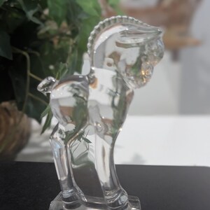May include: A clear glass horse figurine. The horse is standing on a rectangular base. The figurine has a detailed mane and tail. The horse is a decorative item.