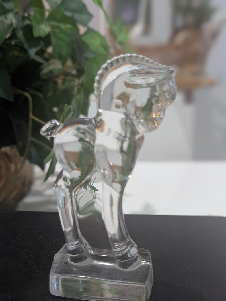 May include: A clear glass horse figurine. The horse is standing on a rectangular base. The figurine has a detailed mane and tail. The horse is a decorative item.
