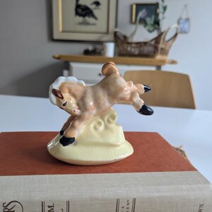 May include: A ceramic figurine of a leaping fawn, featuring tan and white fur with black hooves. The fawn is perched on a yellow cloud base. The figurine is placed on a red book with a beige cover.