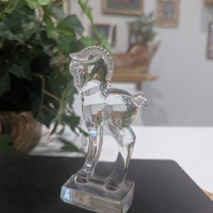 May include: A clear glass horse figurine. The horse is standing on a rectangular base. The figurine has a detailed mane and tail. The horse is transparent, allowing light to pass through.