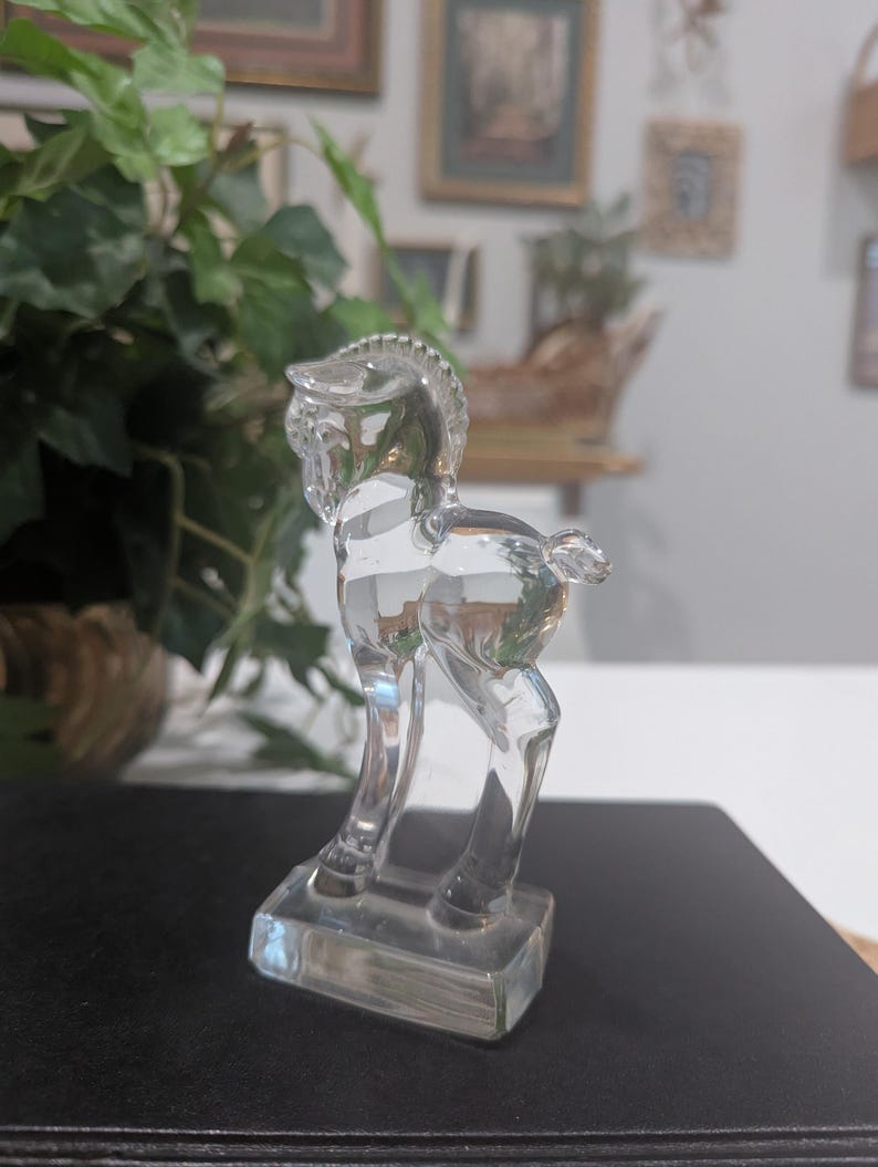 May include: A clear glass horse figurine. The horse is standing on a rectangular base. The figurine has a detailed mane and tail. The horse is transparent, allowing light to pass through.