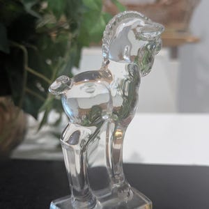 May include: A clear glass figurine of a horse, standing on a rectangular base. The horse is depicted in a stylized, Art Deco design. The figurine is transparent, allowing light to pass through, and is set against a blurred background.