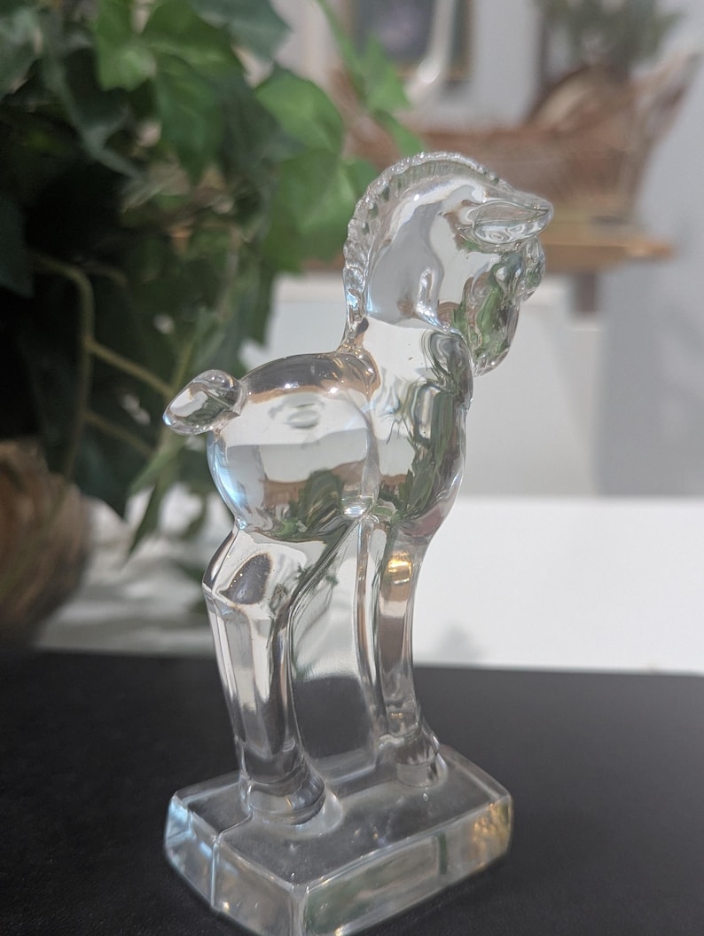 May include: A clear glass figurine of a horse, standing on a rectangular base. The horse is depicted in a stylized, Art Deco design. The figurine is transparent, allowing light to pass through, and is set against a blurred background.