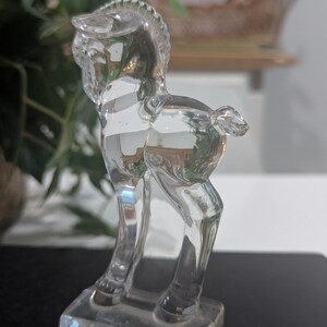May include: A clear glass figurine of a horse, standing on a rectangular base. The horse has a detailed mane and tail, and its body reflects the surrounding light. The figurine is a decorative item.