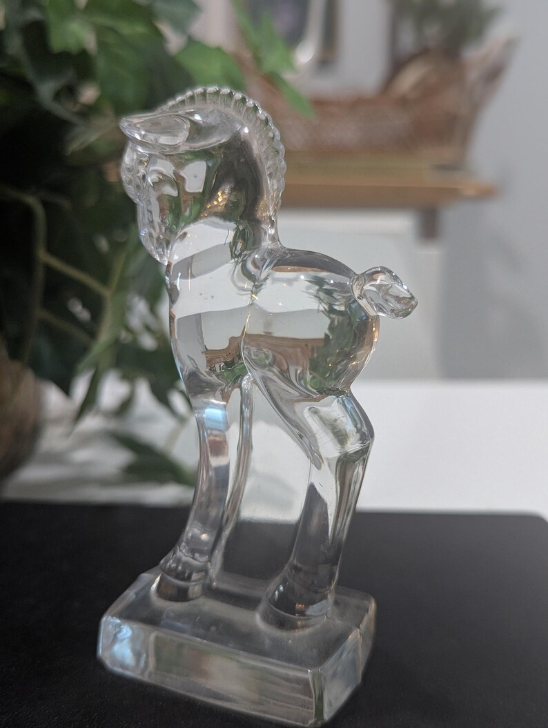 May include: A clear glass figurine of a horse, standing on a rectangular base. The horse has a detailed mane and tail, and its body reflects the surrounding light. The figurine is a decorative item.