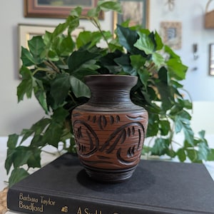 May include: A small, brown vase with carved floral designs and a textured surface. The vase is set on top of a stack of books, with green leafy vines in the background. The vase has a dark brown rim and base.