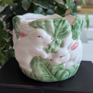 May include: White ceramic planter with a bunny rabbit design. The planter features three white bunnies with pink accents and red eyes, nestled among green leaves. The planter is sitting on a black book.