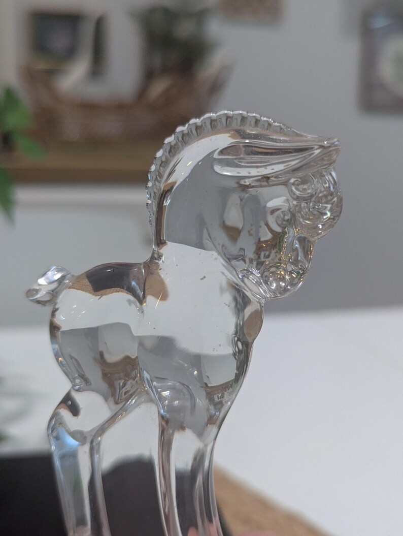 May include: A clear glass figurine of a horse. The horse is standing with its head up and its mane and tail are detailed. The figurine is transparent and reflects light, creating a sparkling effect. The horse is a decorative item.