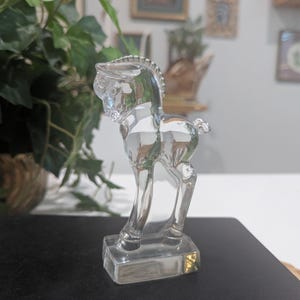 May include: A clear glass horse figurine. The horse is standing on a rectangular base. The figurine has detailed features, including a mane, tail, and defined legs. The horse is transparent, allowing light to pass through.