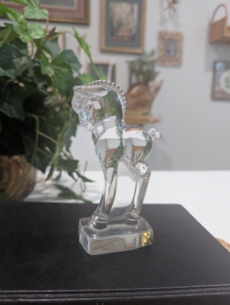 May include: A clear glass horse figurine. The horse is standing on a rectangular base. The figurine has detailed features, including a mane, tail, and defined legs. The horse is transparent, allowing light to pass through.
