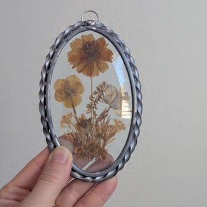 May include: Oval-shaped pressed flower art piece. The frame is silver-colored with a twisted rope design. Inside, dried flowers in shades of brown and yellow are displayed, showcasing a natural aesthetic. A small metal loop is at the top for hanging.