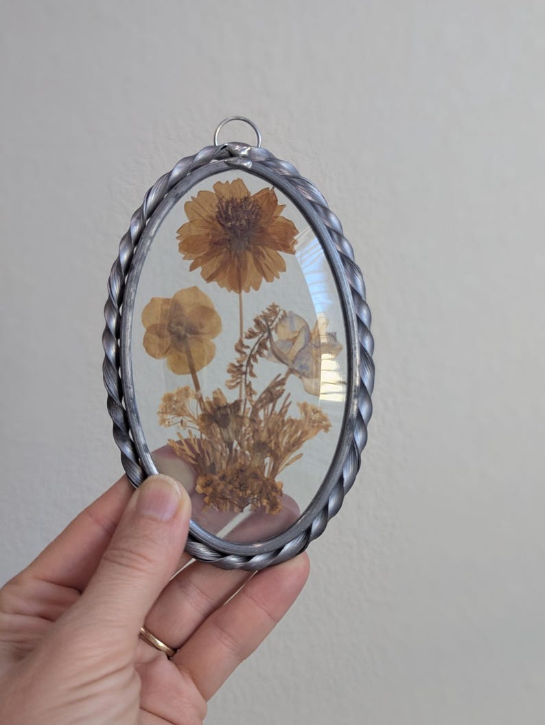 May include: Oval-shaped pressed flower art piece. The frame is silver-colored with a twisted rope design. Inside, dried flowers in shades of brown and yellow are displayed, showcasing a natural aesthetic. A small metal loop is at the top for hanging.