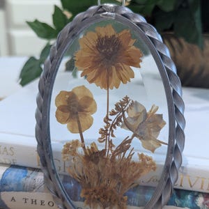 May include: An oval-shaped, silver-toned metal pendant with a clear glass front, showcasing dried flowers in shades of yellow and brown. The pendant has a braided metal border and a small loop at the top for hanging. The flowers are arranged in a natural, botanical style.