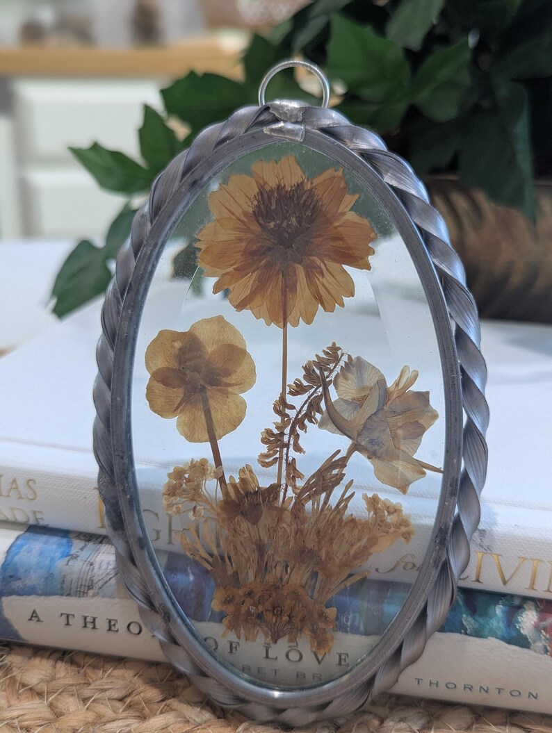 May include: An oval-shaped, silver-toned metal pendant with a clear glass front, showcasing dried flowers in shades of yellow and brown. The pendant has a braided metal border and a small loop at the top for hanging. The flowers are arranged in a natural, botanical style.