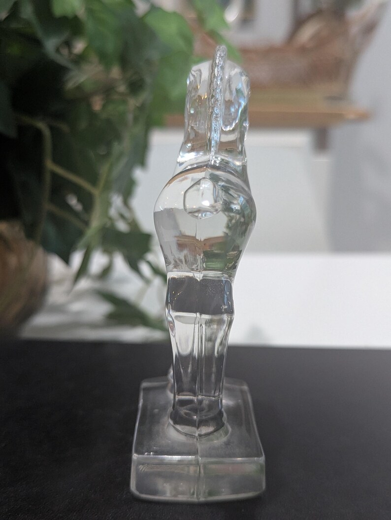 May include: A clear glass horse figurine. The horse is standing on a rectangular base. The figurine is transparent, allowing light to pass through, and is positioned against a dark surface with a blurred background of greenery.