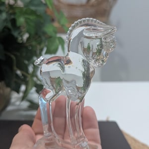 May include: A clear glass horse figurine. The horse is standing on a rectangular base. The figurine has a detailed mane and facial features. The horse is a decorative item.