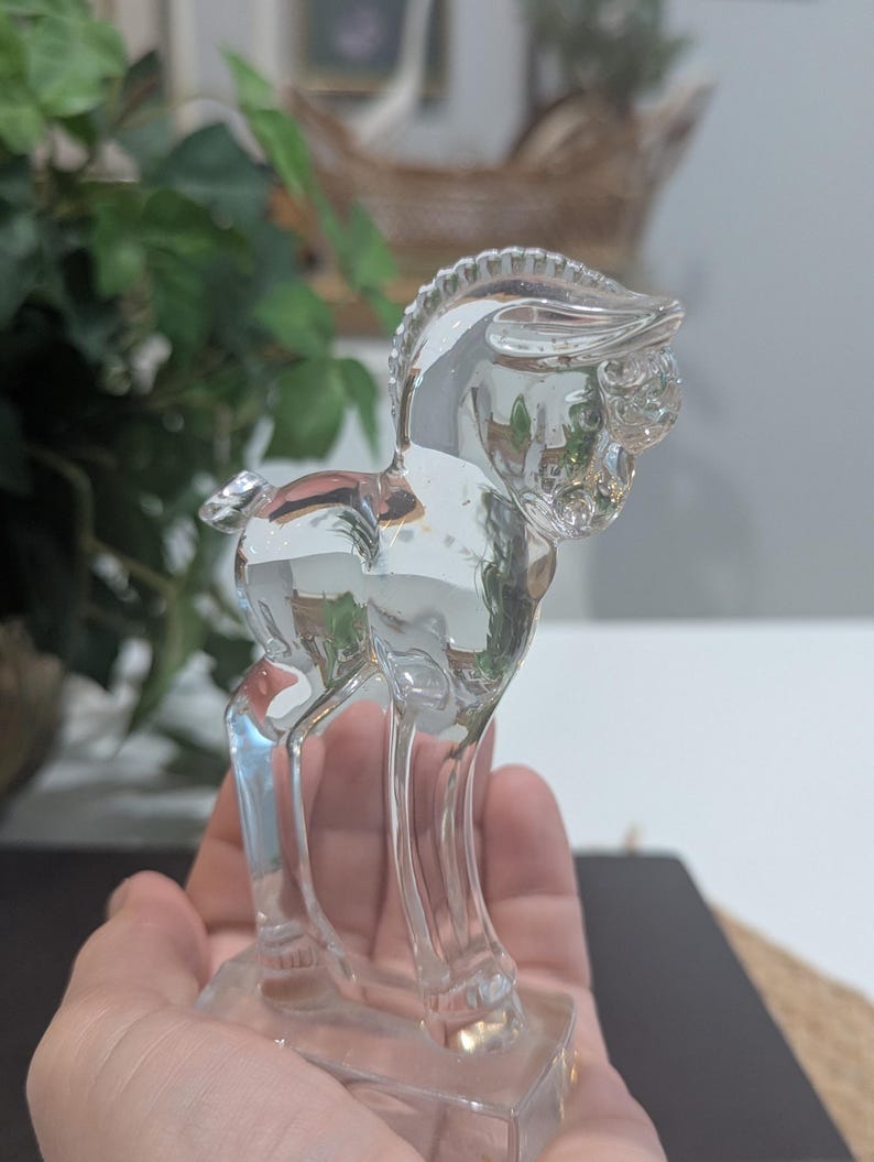May include: A clear glass horse figurine. The horse is standing on a rectangular base. The figurine has a detailed mane and facial features. The horse is a decorative item.