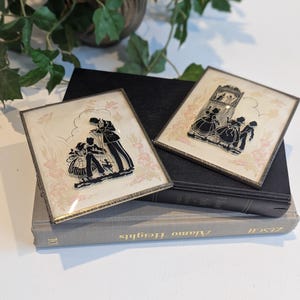 May include: Two framed silhouette art pieces with gold trim, depicting figures in period clothing. The artwork is set against a cream background with a floral pattern. The frames rest on a stack of books, with greenery in the background.