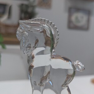May include: A clear glass horse figurine. The horse is depicted in a standing pose, with detailed features including a mane, tail, and defined musculature. The glass is transparent, allowing light to pass through, creating highlights and shadows.