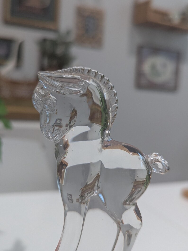 May include: A clear glass horse figurine. The horse is depicted in a standing pose, with detailed features including a mane, tail, and defined musculature. The glass is transparent, allowing light to pass through, creating highlights and shadows.