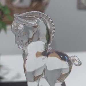 May include: A clear glass figurine of a horse, showcasing intricate details. The horse is posed with its head turned, revealing a detailed mane and tail. The transparent material allows light to pass through, highlighting the form.