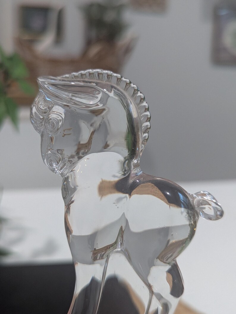May include: A clear glass figurine of a horse, showcasing intricate details. The horse is posed with its head turned, revealing a detailed mane and tail. The transparent material allows light to pass through, highlighting the form.